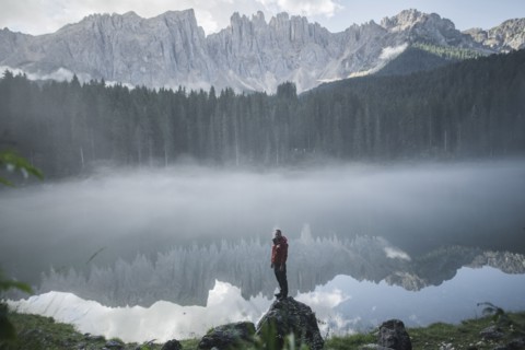 Italy, Carezza, Young man standing on rock at Lago di Carezza in Dolomite Alps at dawn