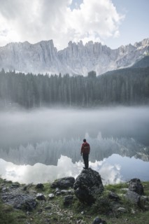 Italy, Carezza, Young man standing on rock at Lago di Carezza in Dolomite Alps at dawn
