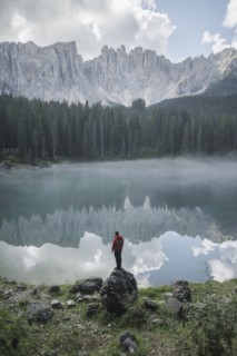 Italy, Carezza, Young man standing on rock at Lago di Carezza in Dolomite Alps at dawn