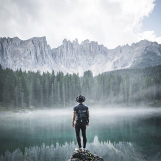Italy, Carezza, Young man standing on rock at Lago di Carezza in Dolomite Alps at dawn