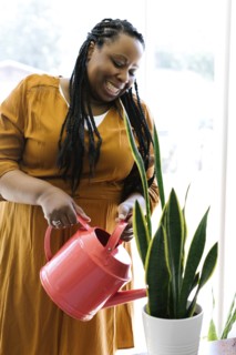 Smiling woman in orange dress watering potted plant at home