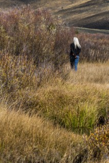 Senior woman with long white hair walking by stream among grass in non urban landscape
