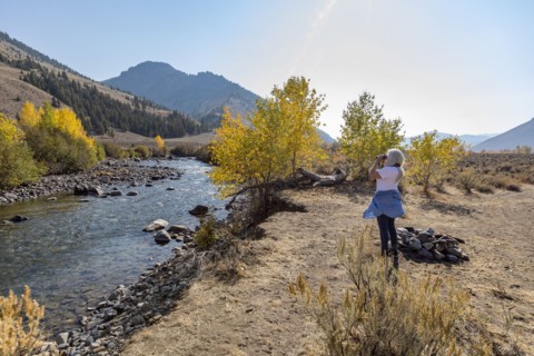 Senior woman looking through binoculars while standing by river in autumn landscape