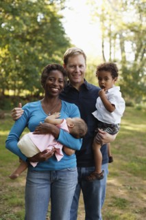 Family outdoors posing for photograph holding preschool boy and baby girl looking at camera smiling