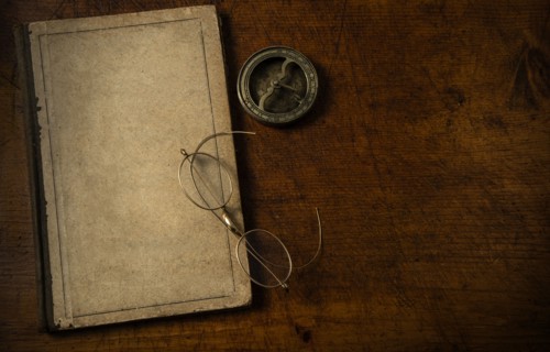 Old book with blank cover resting on old wooden desk top with old eyeglasses and small sundial