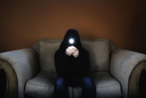 Man in hood sitting on sofa and holding flashlight in front of his face