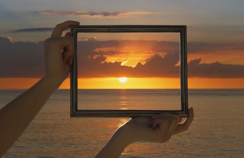 Caucasian woman holding frame over dramatic sunset sky and ocean