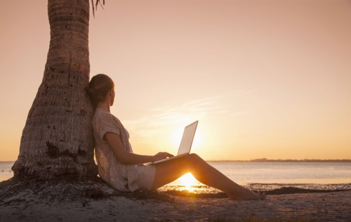 Caucasian woman with laptop leaning on palm tree at sunset