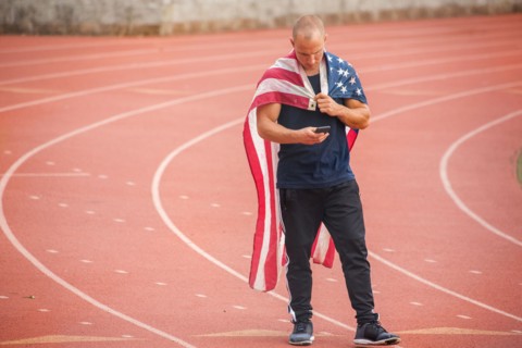 Caucasian man wearing American flag as cape on track texting on cell phone