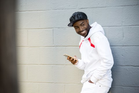 Smiling Black man leaning on wall holding cell phone
