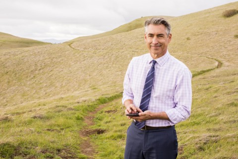 Smiling Caucasian businessman standing in grass holding cell phone