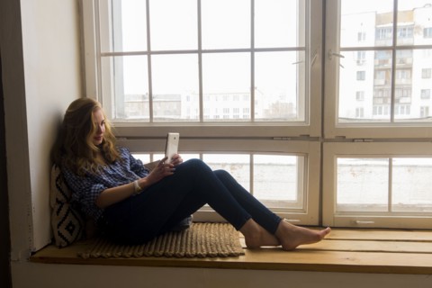 Caucasian woman sitting on window sill reading digital tablet