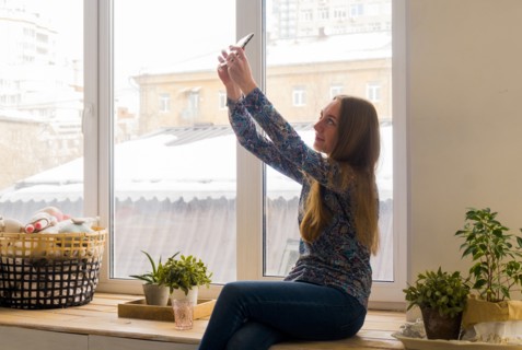 Caucasian woman sitting at window posing for cell phone selfie