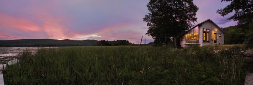Panoramic shot of an illuminated cabin with clapboard siding by lake at dusk