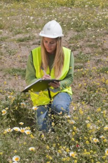Hispanic worker in hard-hat kneeling in field with clipboard