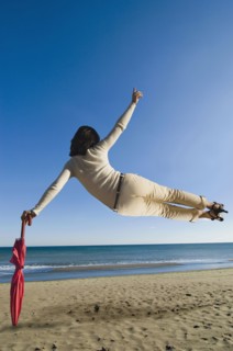 Woman jumping mid air on beach leaning on umbrella rear view