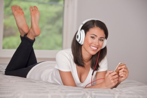 Hispanic woman laying on bed listening to cell phone with headphones
