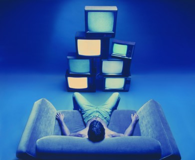 Man sitting on loveseat watching stack of televisions