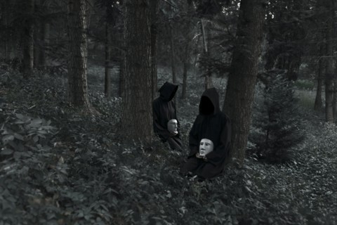 People wearing black robes and holding white masks sitting in forest