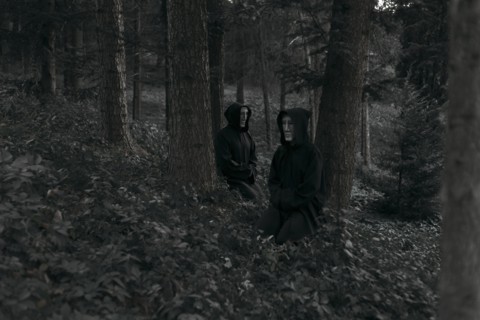 People wearing black robes and white masks sitting in forest