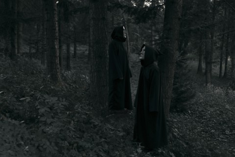 People wearing black robes and white masks standing in forest