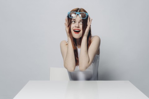Happy Caucasian woman sitting at table lifting blue eyeglasses