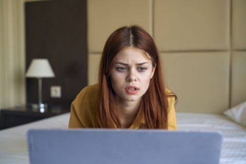 Frustrated Caucasian woman laying on bed using laptop