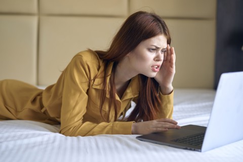 Frustrated Caucasian woman laying on bed using laptop