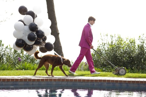 Caucasian man in pink suit mowing lawn being followed by dog