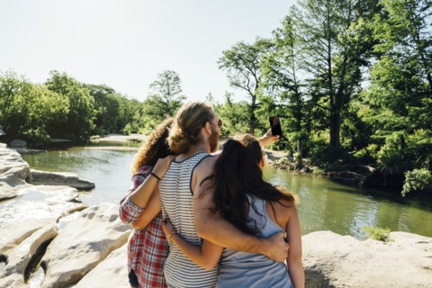 Friends posing for cell phone selfie on rocks near river
