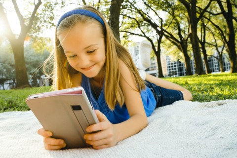 Smiling Caucasian girl laying on blanket in park reading digital tablet