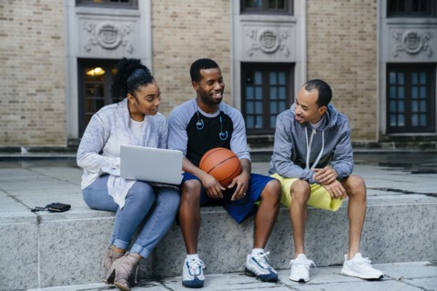 Black friends sitting on concrete with basketball and laptop
