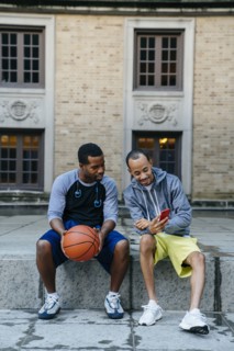 Black men sitting on concrete with basketball and texting on cell phone