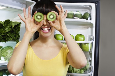 Mixed race woman covering eyes with kiwi slices near refrigerator