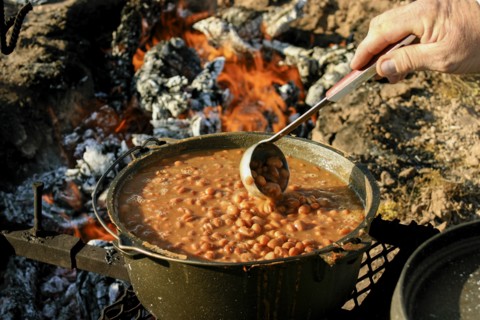 Hand of man stirring beans with ladle on campfire