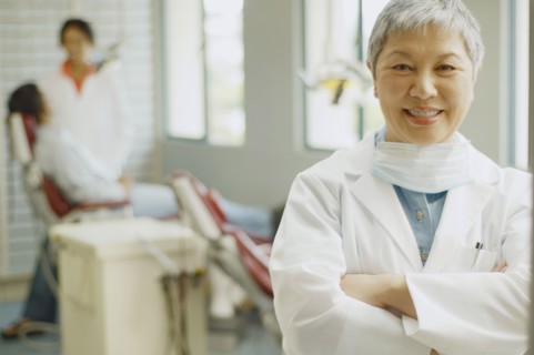 Senior Asian female dentist with assistant and patient in background
