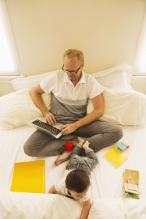 Father using laptop and playing with son on bed