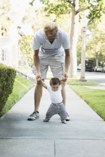 Father helping baby son walk on sidewalk