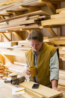 Caucasian carpenter drinking coffee and using cell phone in workshop