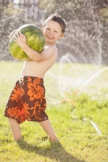Caucasian boy carrying watermelon through sprinkler