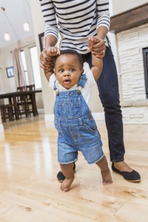 Black mother helping baby son walk on living room floor