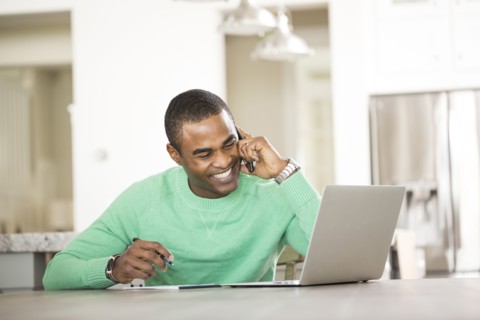 Black man sitting at table with laptop talking on cell phone