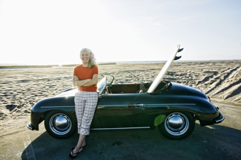 Older Caucasian woman leaning on convertible car with surfboard on beach
