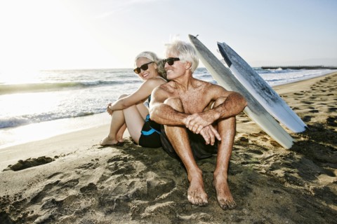 Older Caucasian couple sitting on beach with surfboards