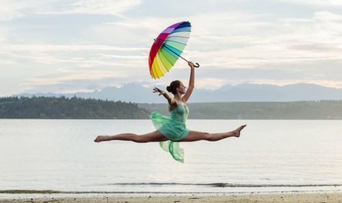 Caucasian ballerina jumping with multicolor umbrella on beach