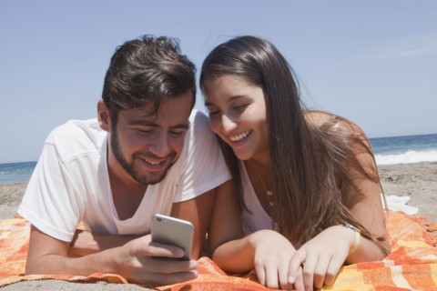Hispanic couple laying on blanket at beach texting on cell phone