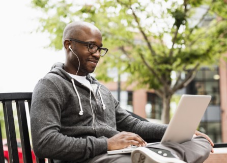 Black man using laptop and earphones on city bench