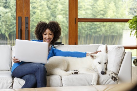 Smiling Black woman petting white dog on sofa using laptop