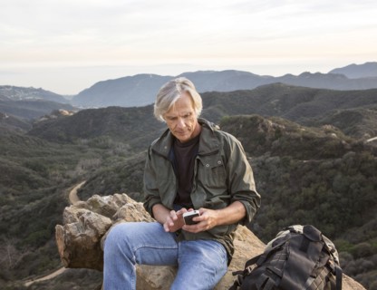 Older Caucasian man using cell phone on rocky hilltop