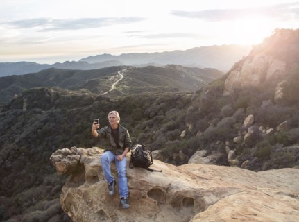 Older Caucasian man taking cell phone photograph on rocky hilltop